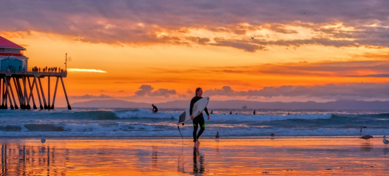 Surfer walking along the beach at sunset with vibrant orange skies and pier in background.