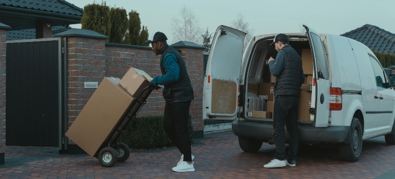 Two professional movers unloading boxes from a white van in a residential neighborhood driveway.