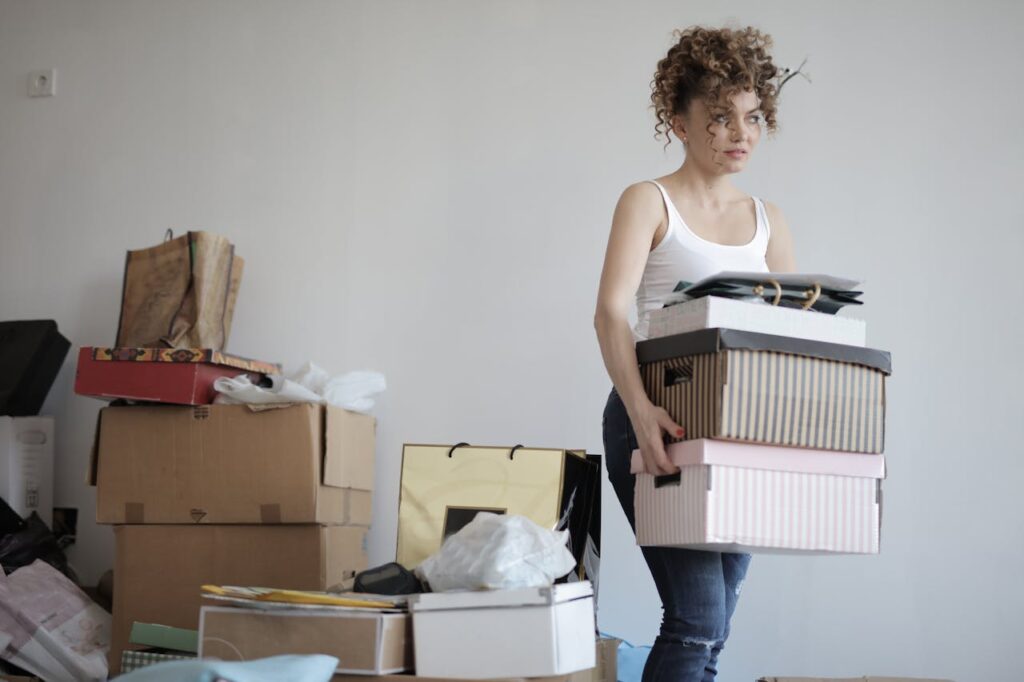 A person in a white tank top carries stacked boxes amid a cluttered room filled with cardboard and paper items.