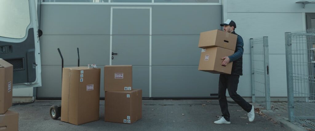 Person carrying stacked cardboard boxes outside a warehouse with a delivery van nearby, representing warehouse moving in LA.