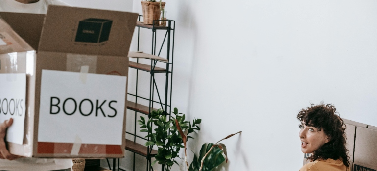 Man holding a labeled box of books while woman sits nearby, smiling during move-in.