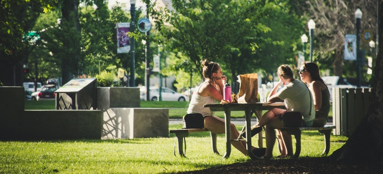 Friends enjoy snacks at a sunny park table, laughing and chatting in a lively outdoor setting.