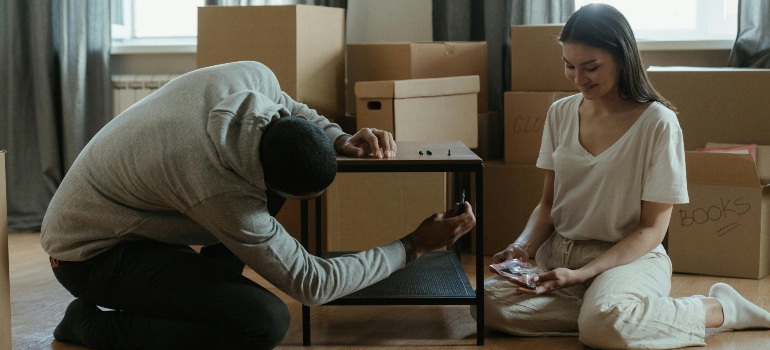 Couple assembling a small table while surrounded by moving boxes.