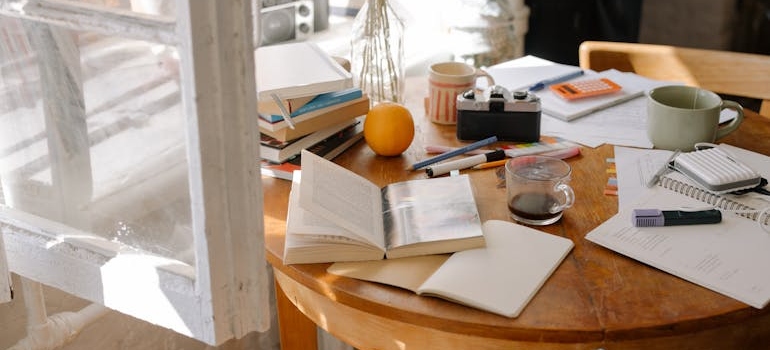 Wooden table covered with books, notes, and personal items near a window in a recently packed home.