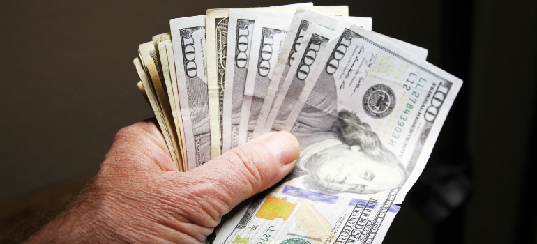 A hand holds a thick stack of hundred-dollar bills, symbolizing savings, expenses, and the Things to Know Before Moving to Glendale.