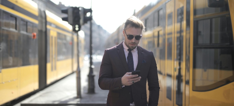 Stylish man in a suit checks his phone near bright yellow trams on a city street.