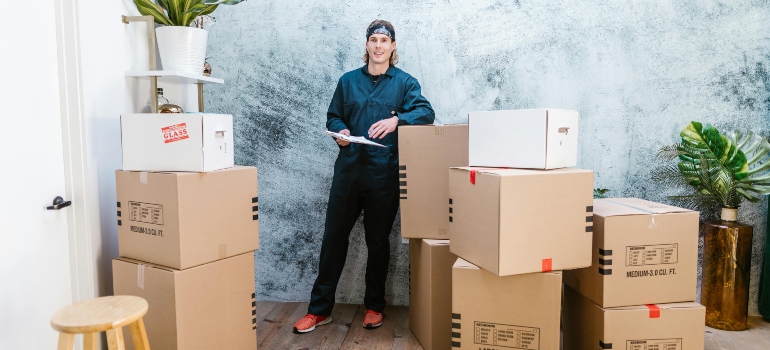 Mover in uniform standing confidently with a clipboard surrounded by stacked moving boxes.