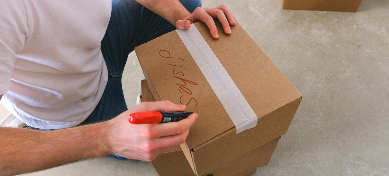 Person labeling a cardboard box 'Dishes' with a red marker while preparing for a move.